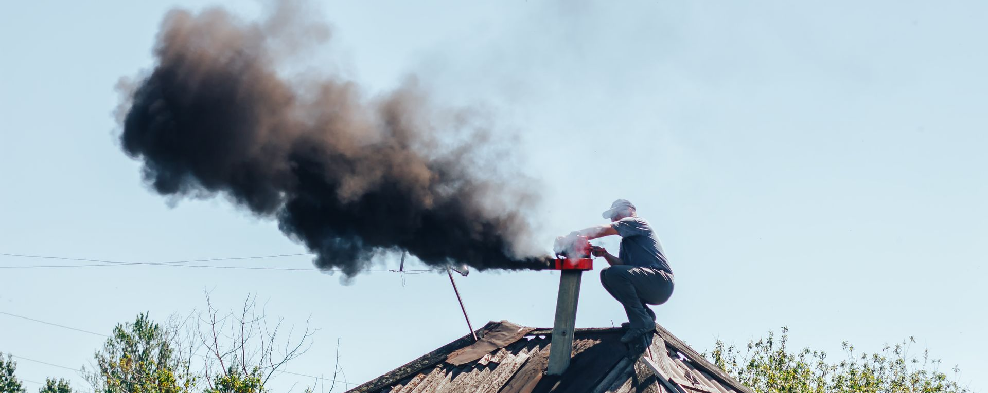 Une personne est accroupie sur un toit près d'une cheminée d'où s'échappe un épais panache de fumée noire sur un ciel bleu.