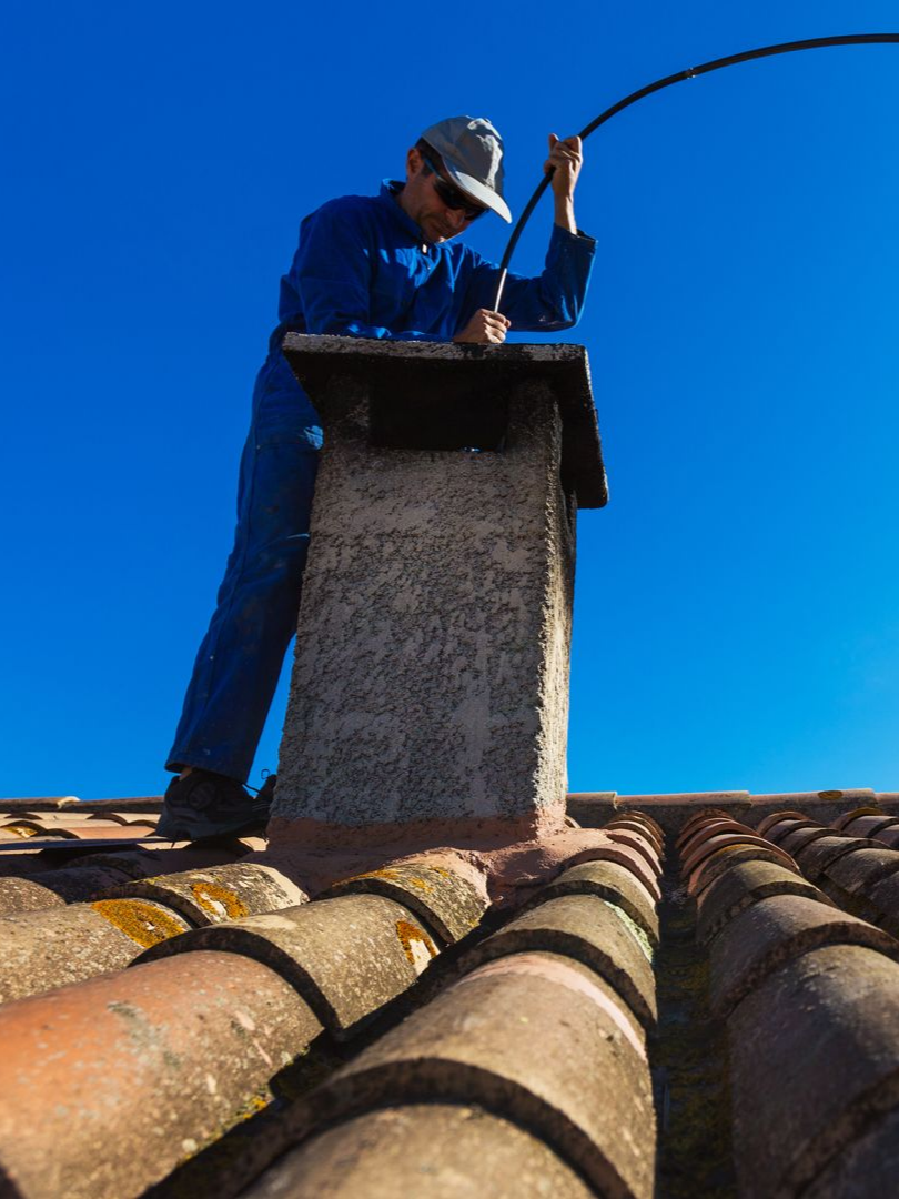 Ramoneur sur un toit, travaillant sur une cheminée avec un long outil flexible, sur fond de ciel bleu clair.
