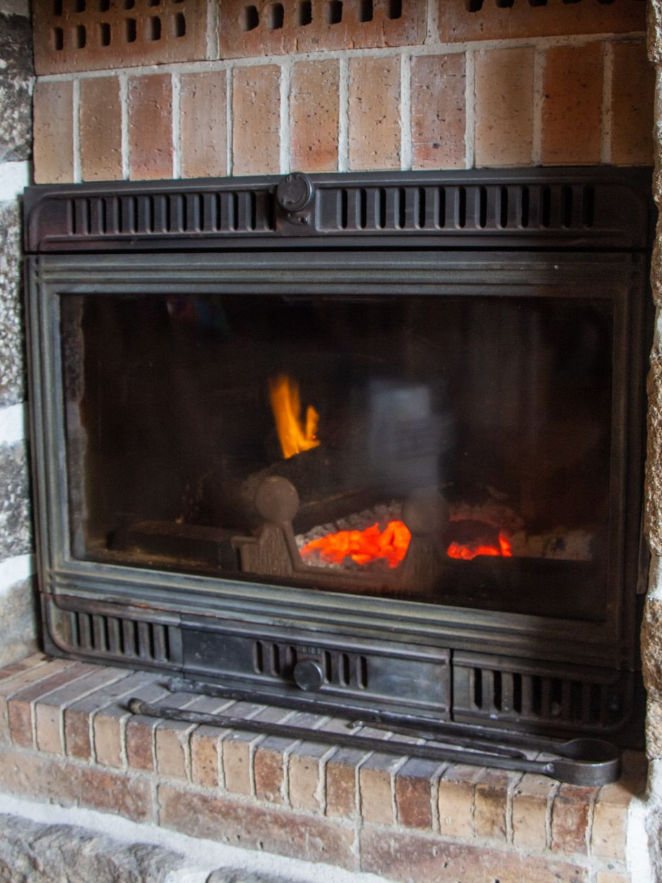 Cheminée avec bûches incandescentes, visible à travers une porte vitrée encadrée de briques.