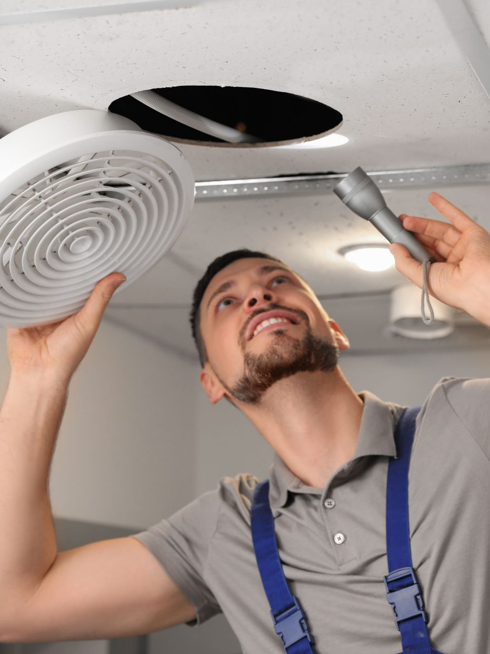 Un homme répare un ventilateur de plafond, lampe torche à la main, inspectant le trou.