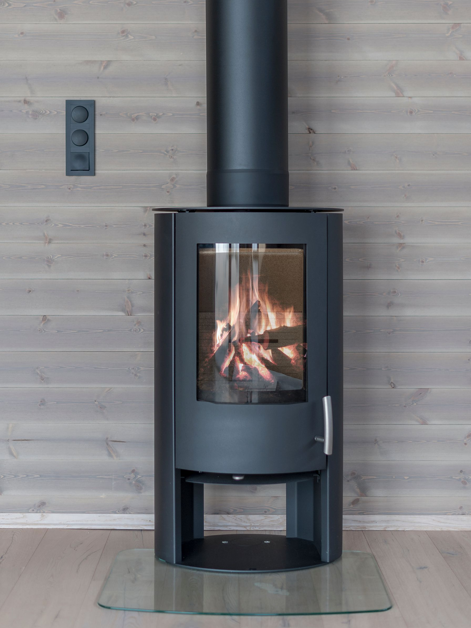 Cheminée noire avec bûches incandescentes, conduit noir, sur une vitre, contre un mur en bois gris.