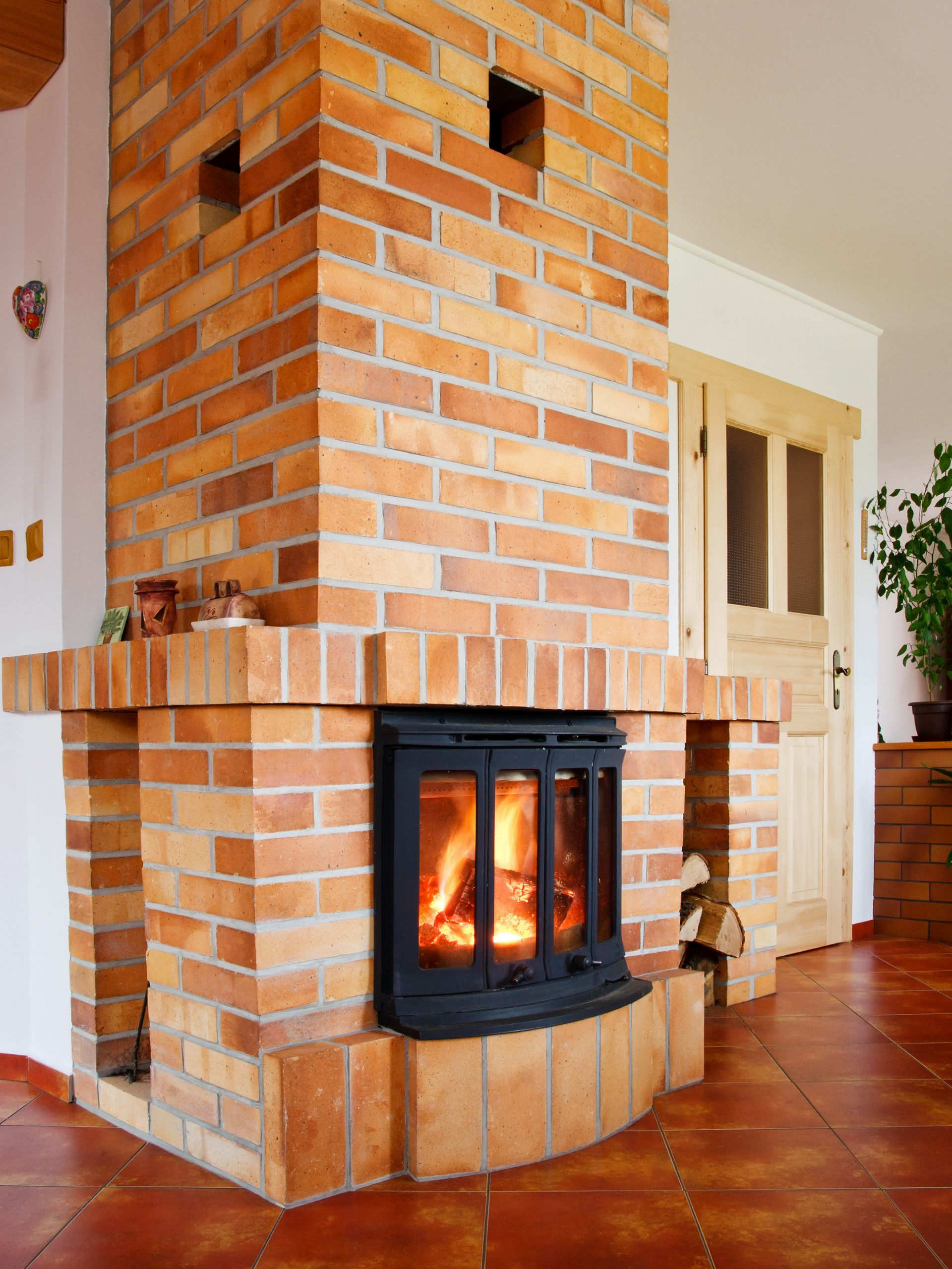 Cheminée en briques avec feu allumé, encastrée dans un angle. Porte en bois et sol carrelé visibles.