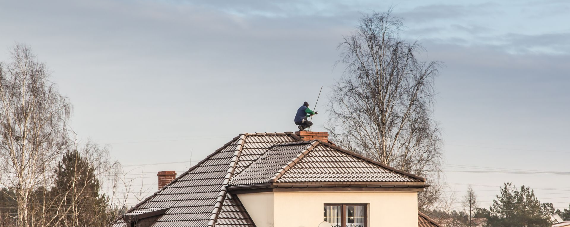 Personne sur une cheminée de toit ; toit enneigé, arbres dénudés, ciel couvert.