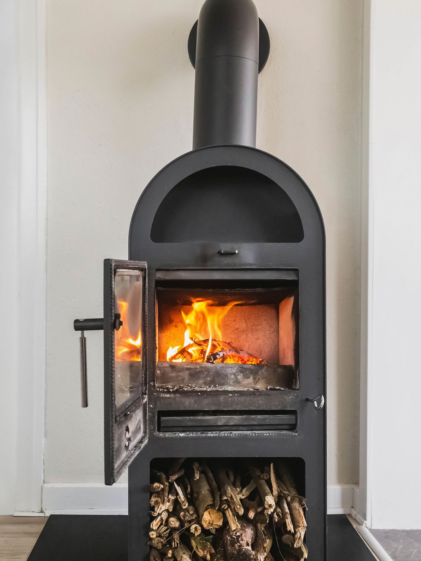 Poêle à bois noir avec porte ouverte, feu qui brûle à l'intérieur. Des bûches sont stockées en dessous.