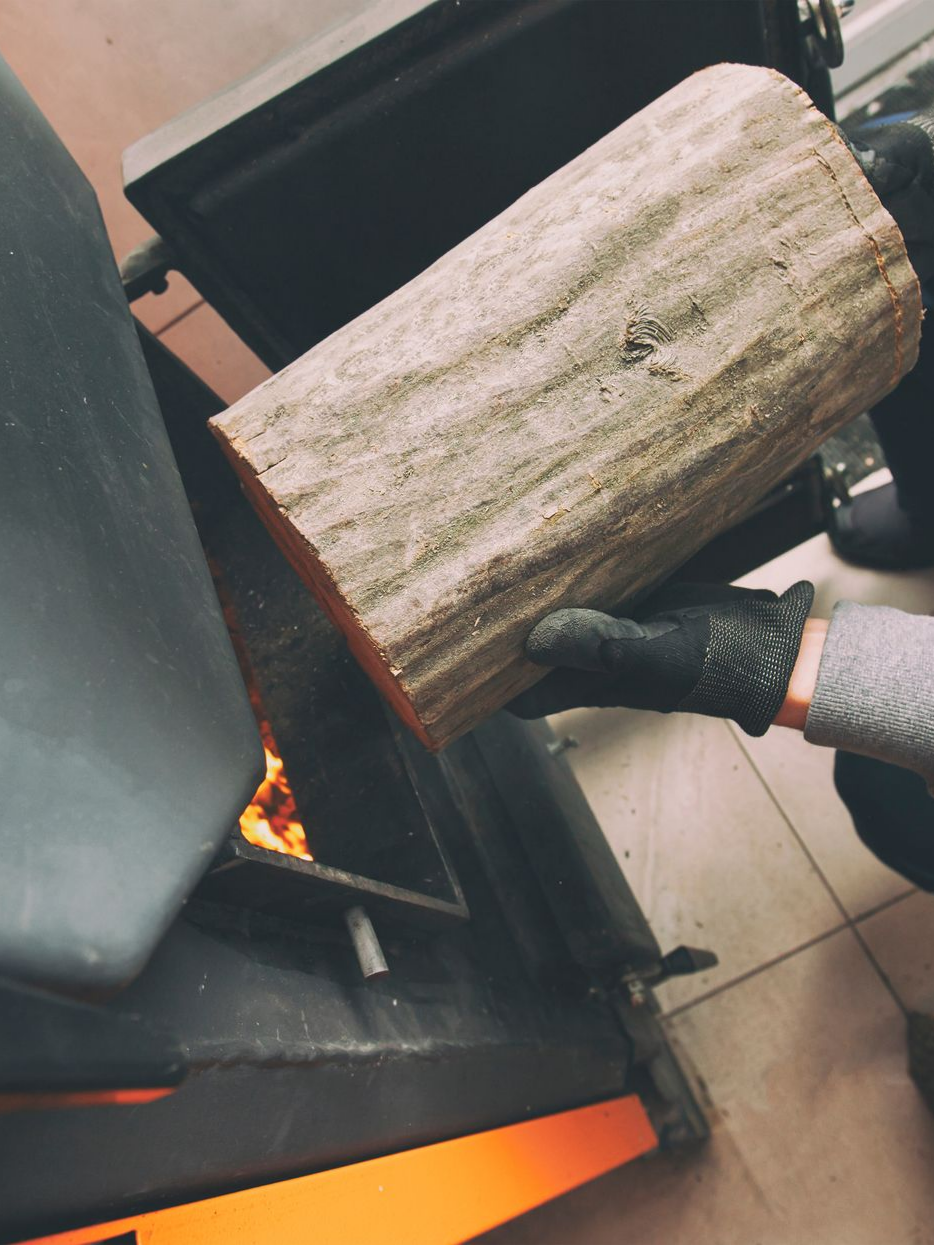 Une personne portant des gants place une bûche dans un poêle à bois dont on peut voir les flammes à l'intérieur.