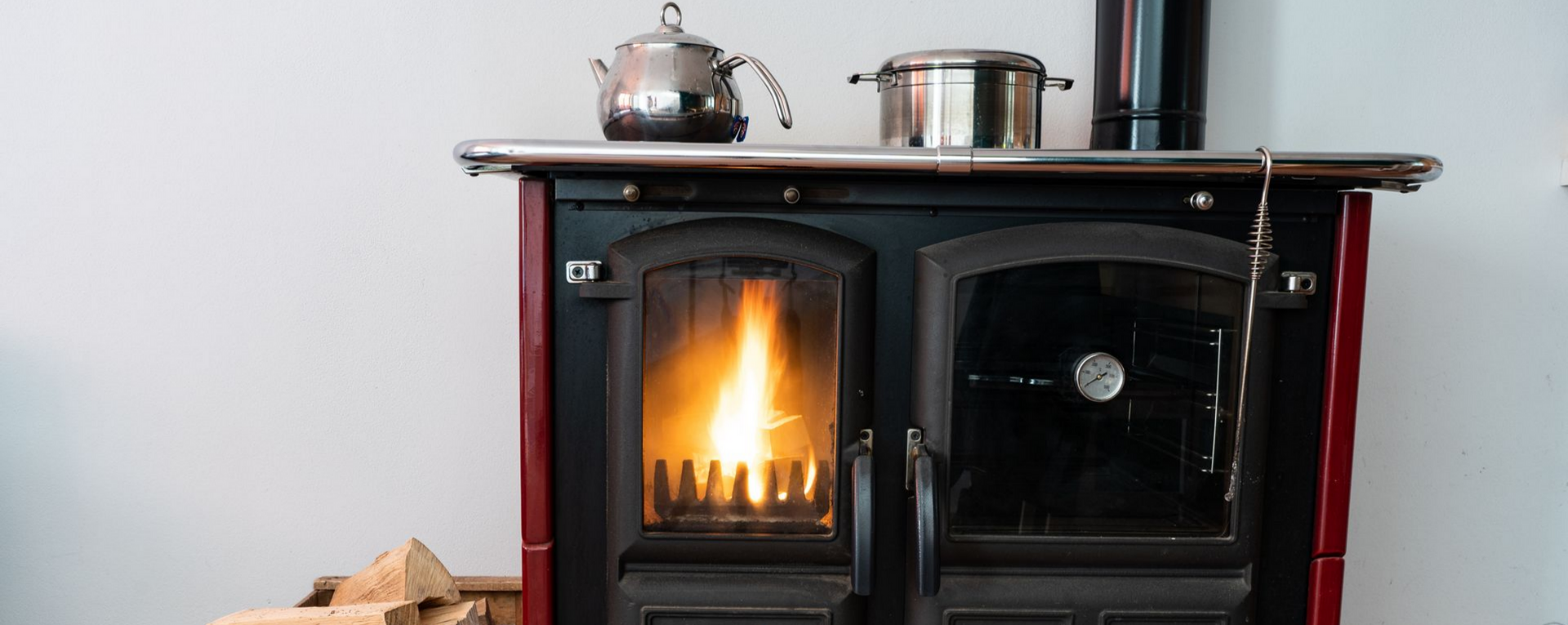 Un poêle à bois allumé avec une théière et une casserole dessus ; du bois de chauffage empilé en dessous.