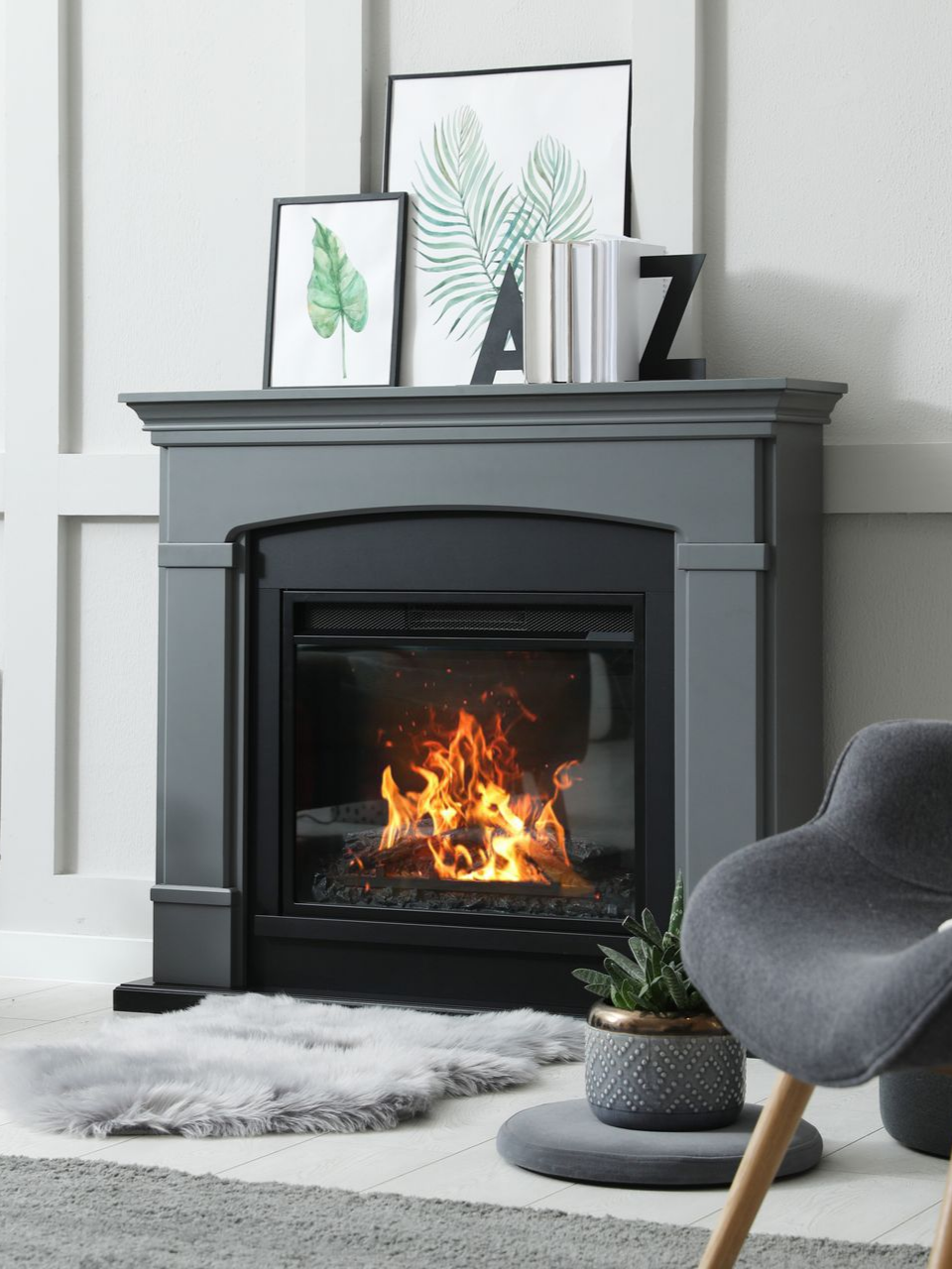 Cheminée grise aux flammes crépitantes, ornée d'œuvres d'art et de livres. Un fauteuil gris se trouve à proximité.