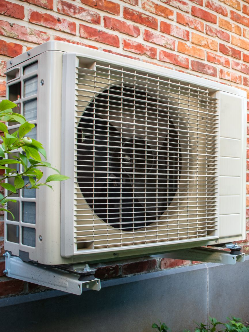 Unité de climatisation fixée sur un mur en briques, avec une grille métallique recouvrant un ventilateur.