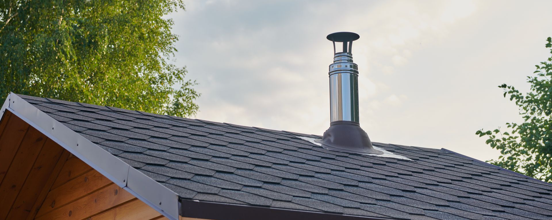Cheminée sur un toit de bardeaux bruns, arbres en arrière-plan. Ciel nuageux.