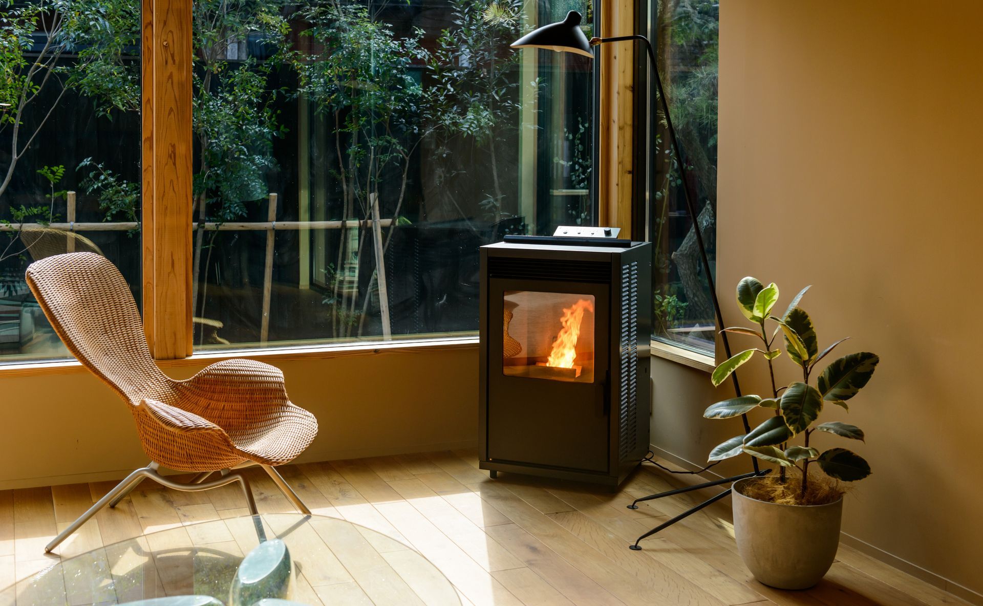 Pièce confortable avec poêle allumé, fauteuil en osier près de la fenêtre, plante d'intérieur et lampe.