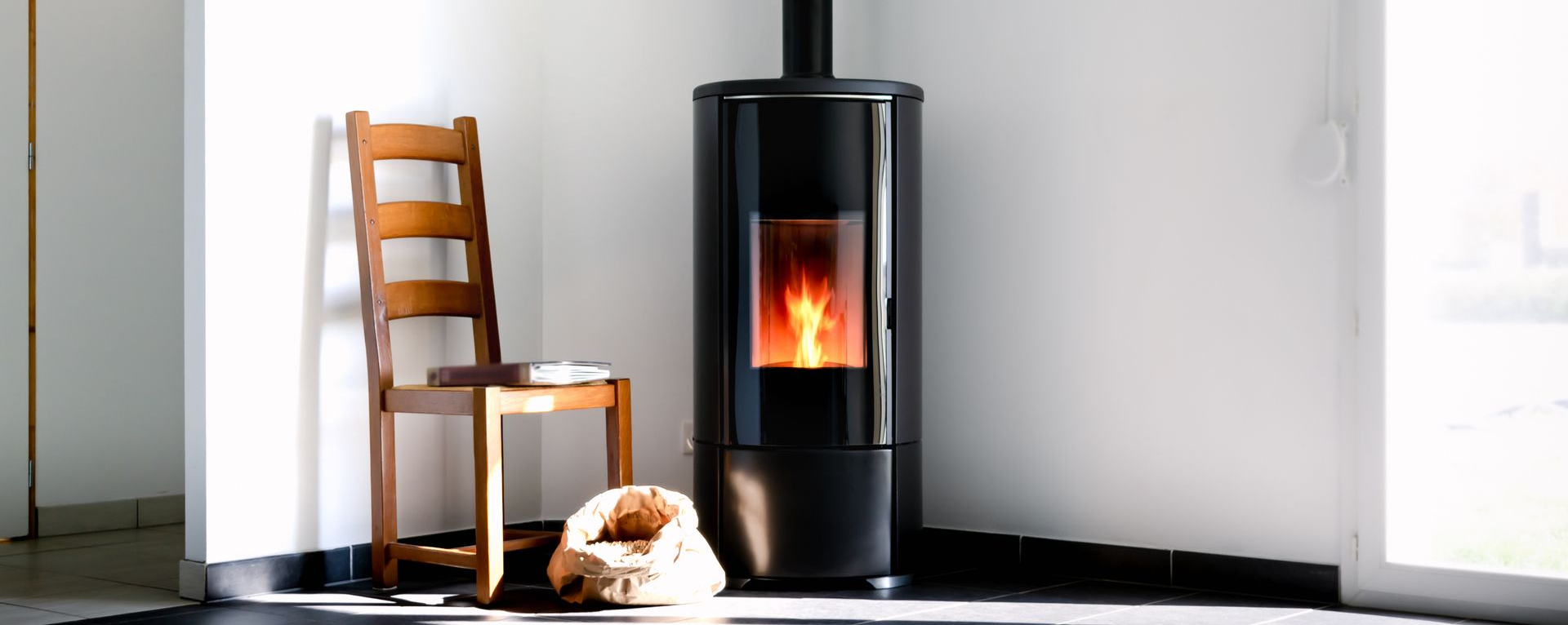 Un poêle à bois noir, avec un feu allumé, se trouve dans un coin de la pièce, à côté d'une chaise en bois.