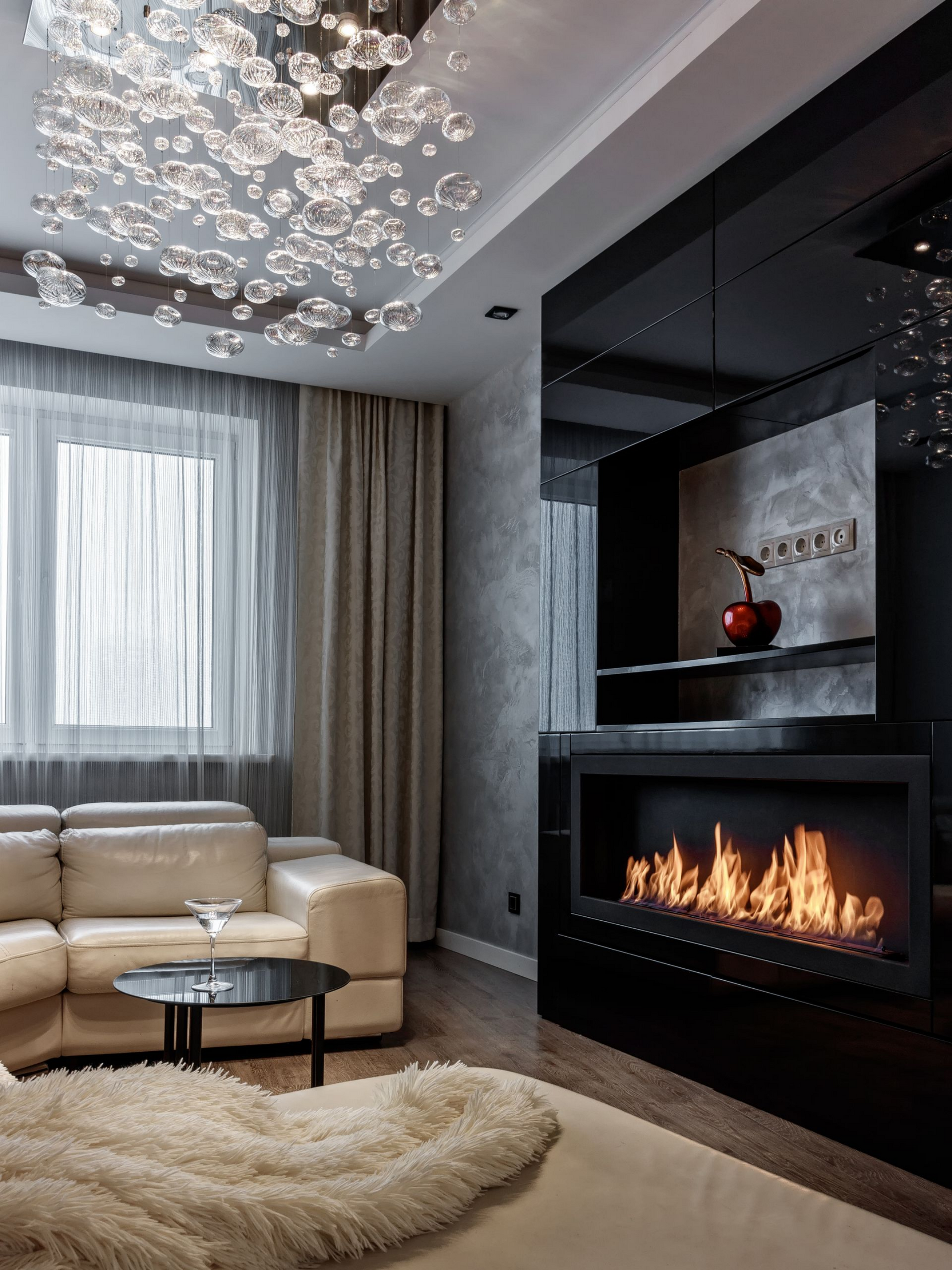 Salon moderne avec cheminée, canapé beige, voilages et lustre à bulles.