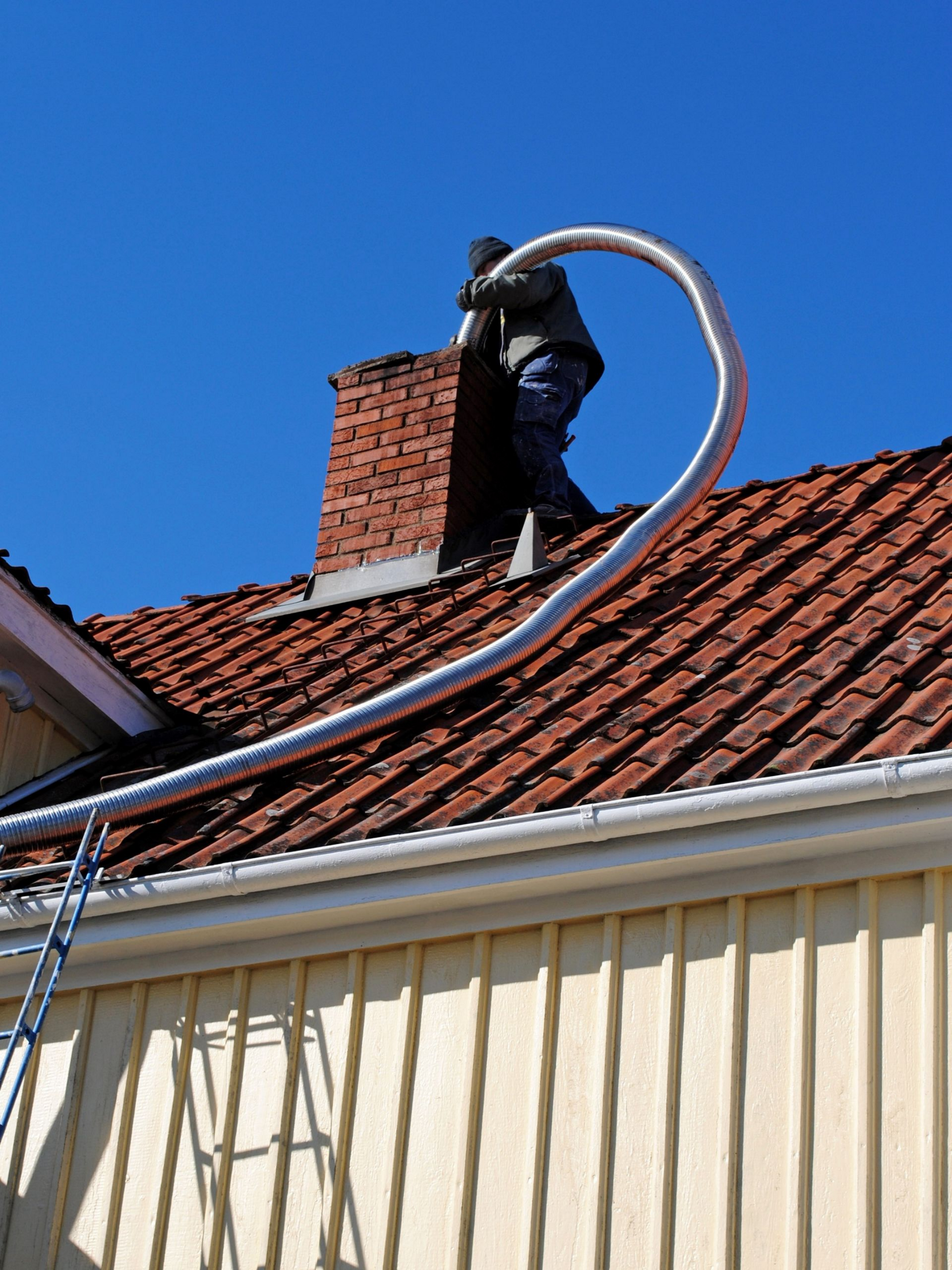 Une personne sur un toit nettoie une cheminée en briques avec un long tuyau flexible.