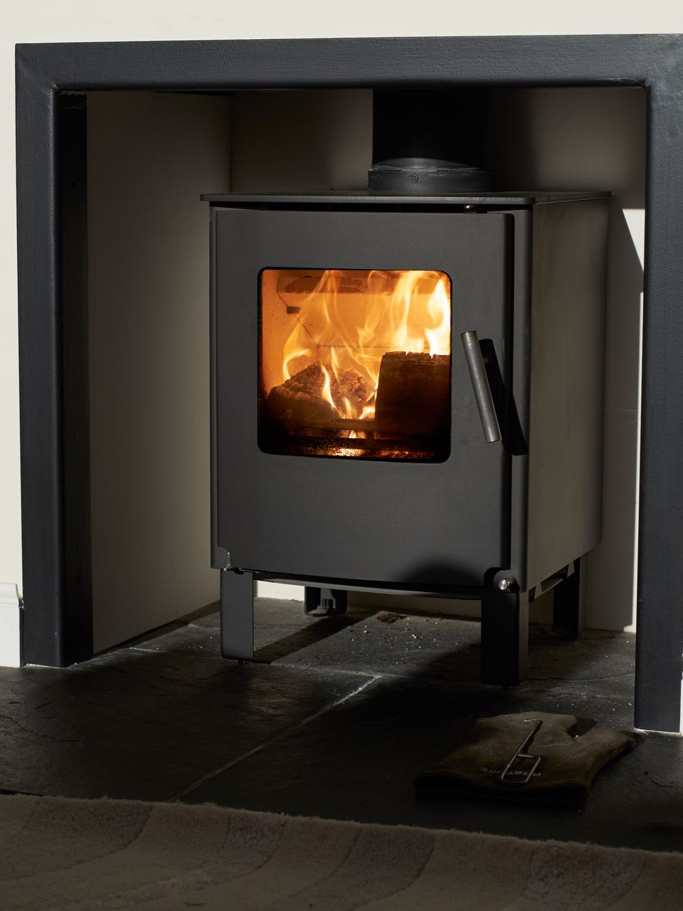 Poêle à bois noir avec des flammes dans une cheminée sombre. Un tapis est posé au sol devant.