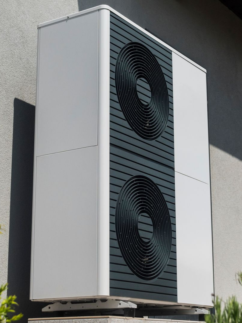 Unité de pompe à chaleur blanche et gris foncé fixée sur un mur gris. Deux ventilateurs circulaires noirs sont visibles.