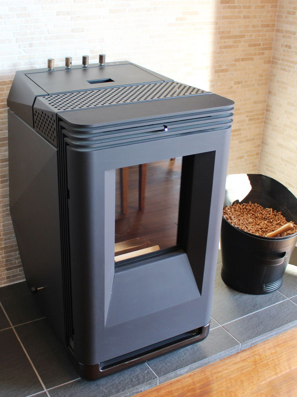 Poêle à granulés noir avec vitre, à côté d'un seau de granulés, sur une surface carrelée.