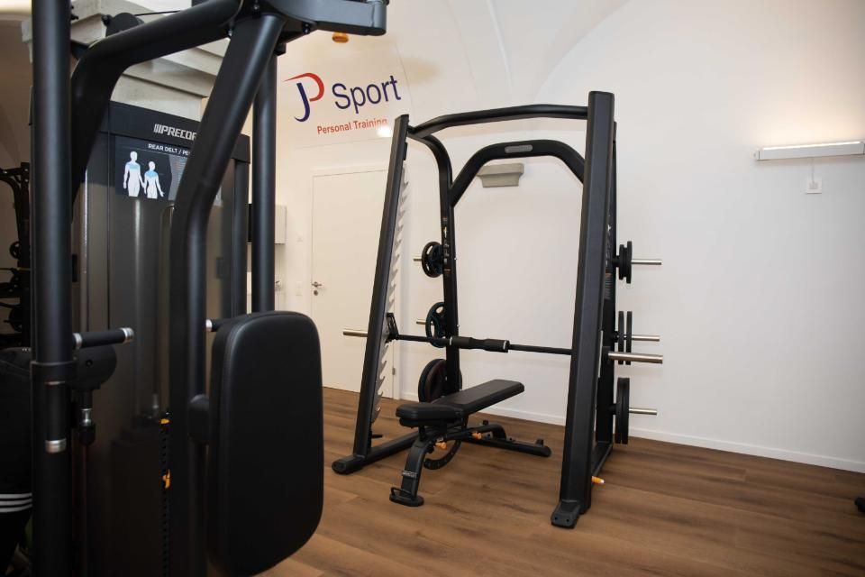 Innenansicht eines Fitnessstudios mit Krafttrainingsgeräten und einer Smith-Maschine vor einer weissen Wand.