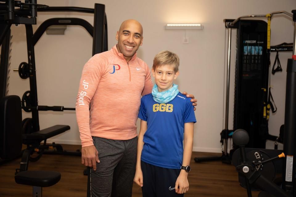 Mann und Junge posieren in einer Turnhalle; der Mann trägt Orange, der Junge Blau, beide lächeln.