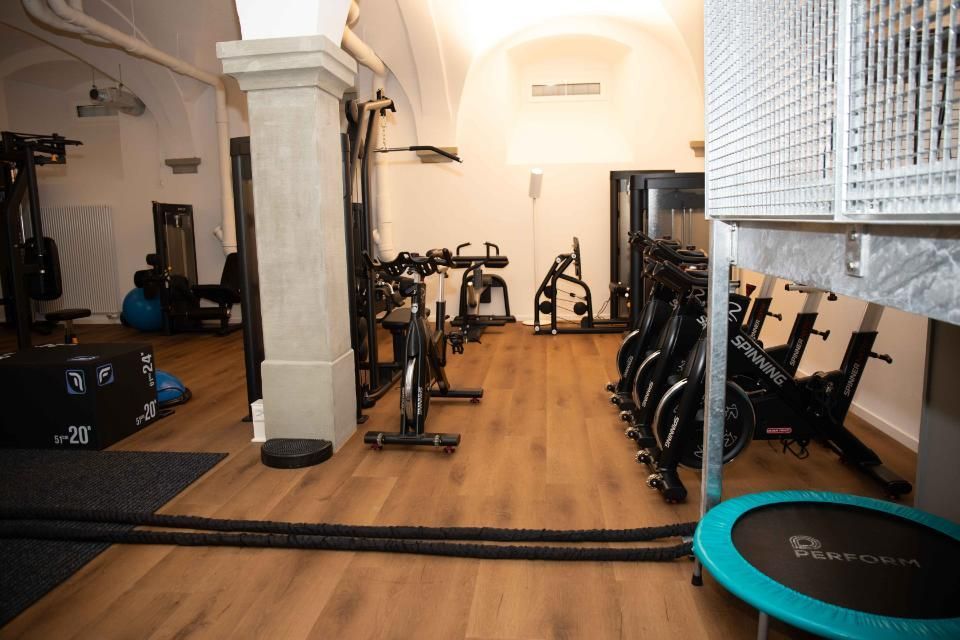 Fitnessstudio-Interieur mit Trainingsgeräten, darunter Fahrräder, Seile und Gewichte. Holzboden, neutrale Wände.