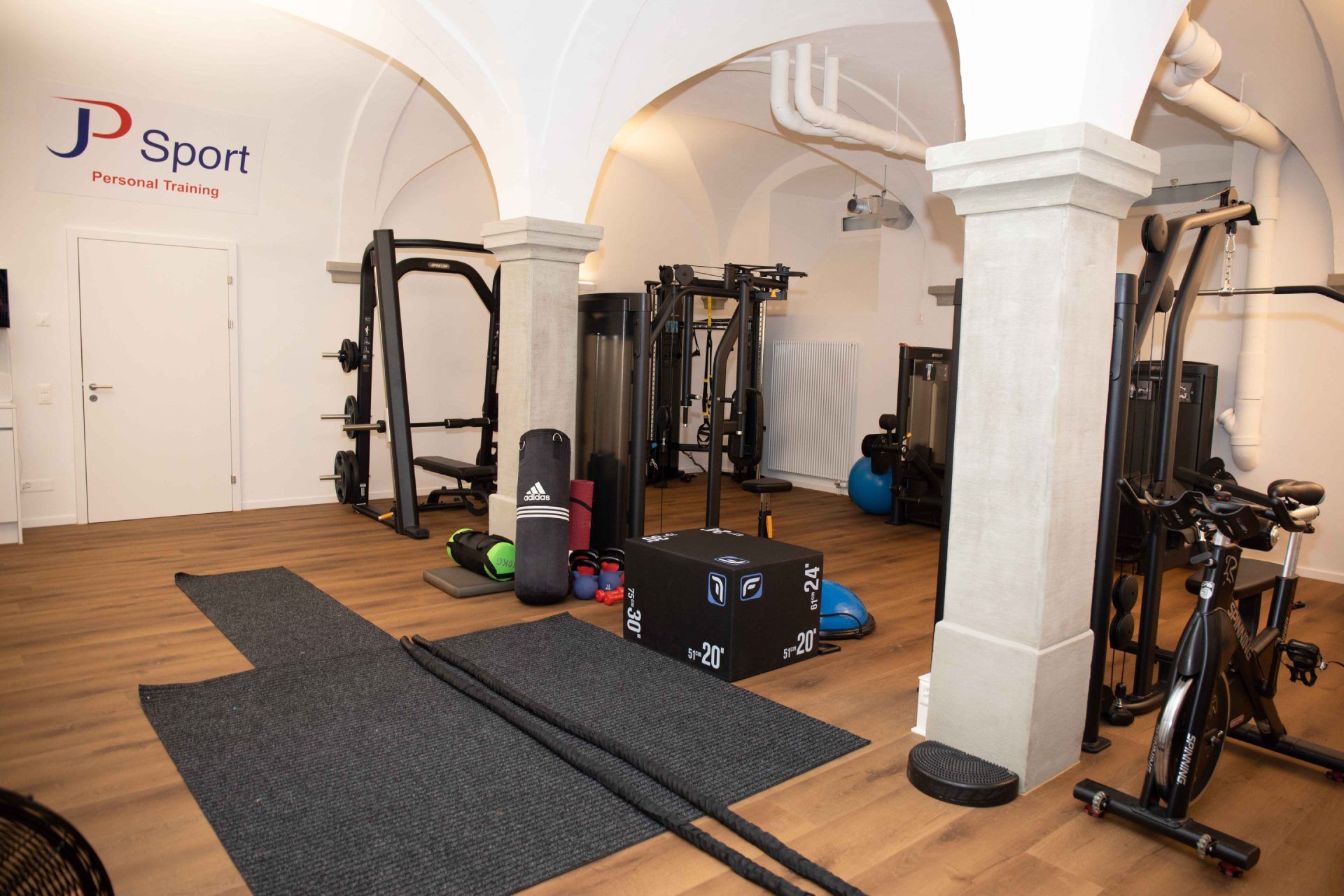 Innenbereich einer Turnhalle mit Trainingsgeräten wie Hantelständern, Fahrrädern und Matten unter gewölbten Decken.