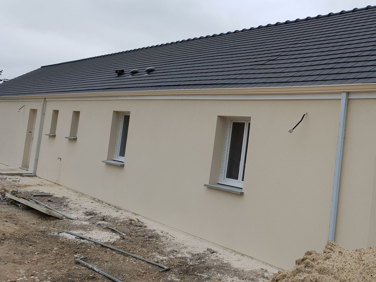 Une maison de plain-pied en construction, avec des murs beiges, des encadrements de fenêtres blancs et un toit de tuiles gris foncé.