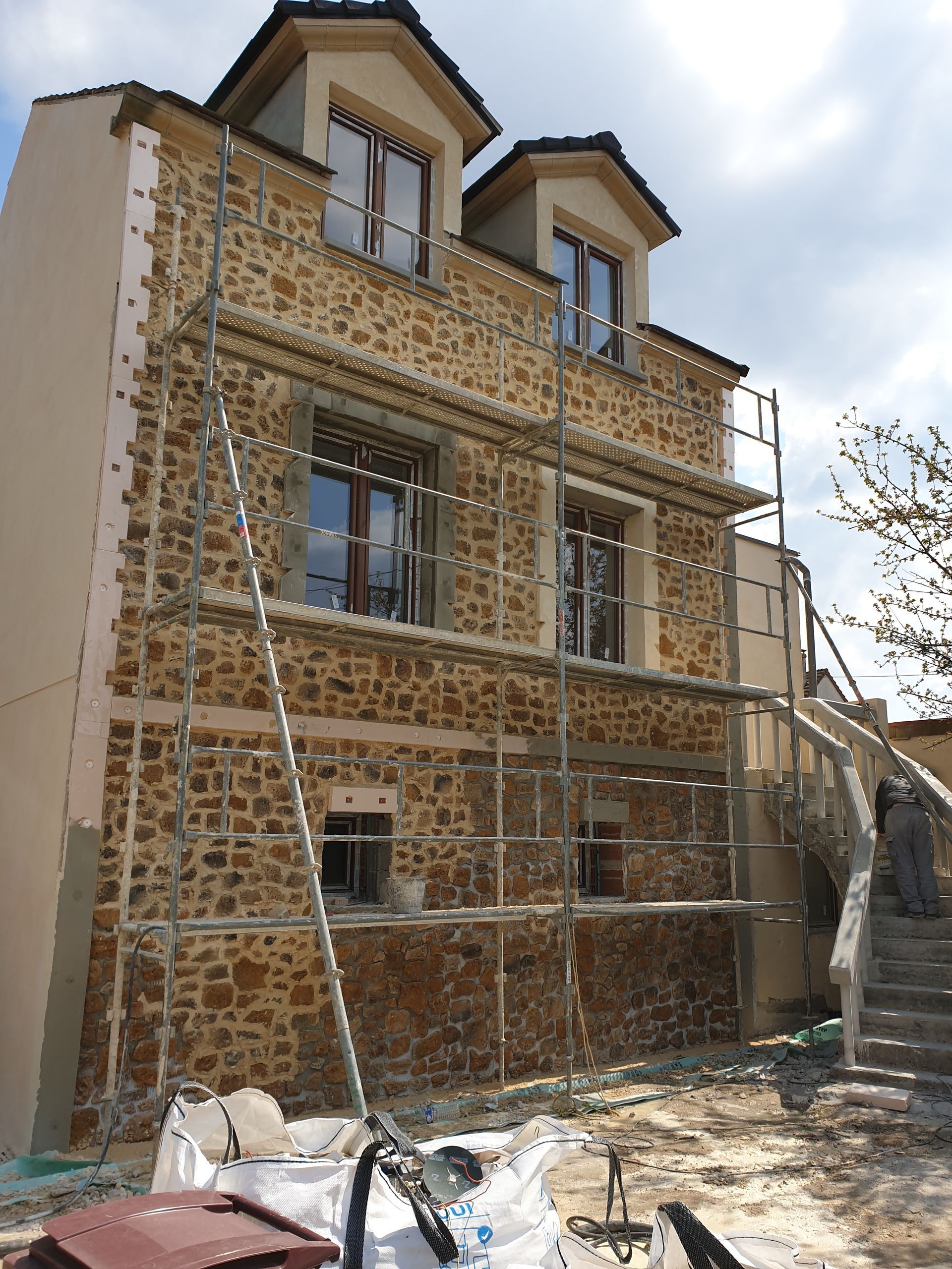 Une maison en pierre de trois étages en cours de rénovation, entourée d'échafaudages métalliques, avec des lucarnes et un escalier latéral.
