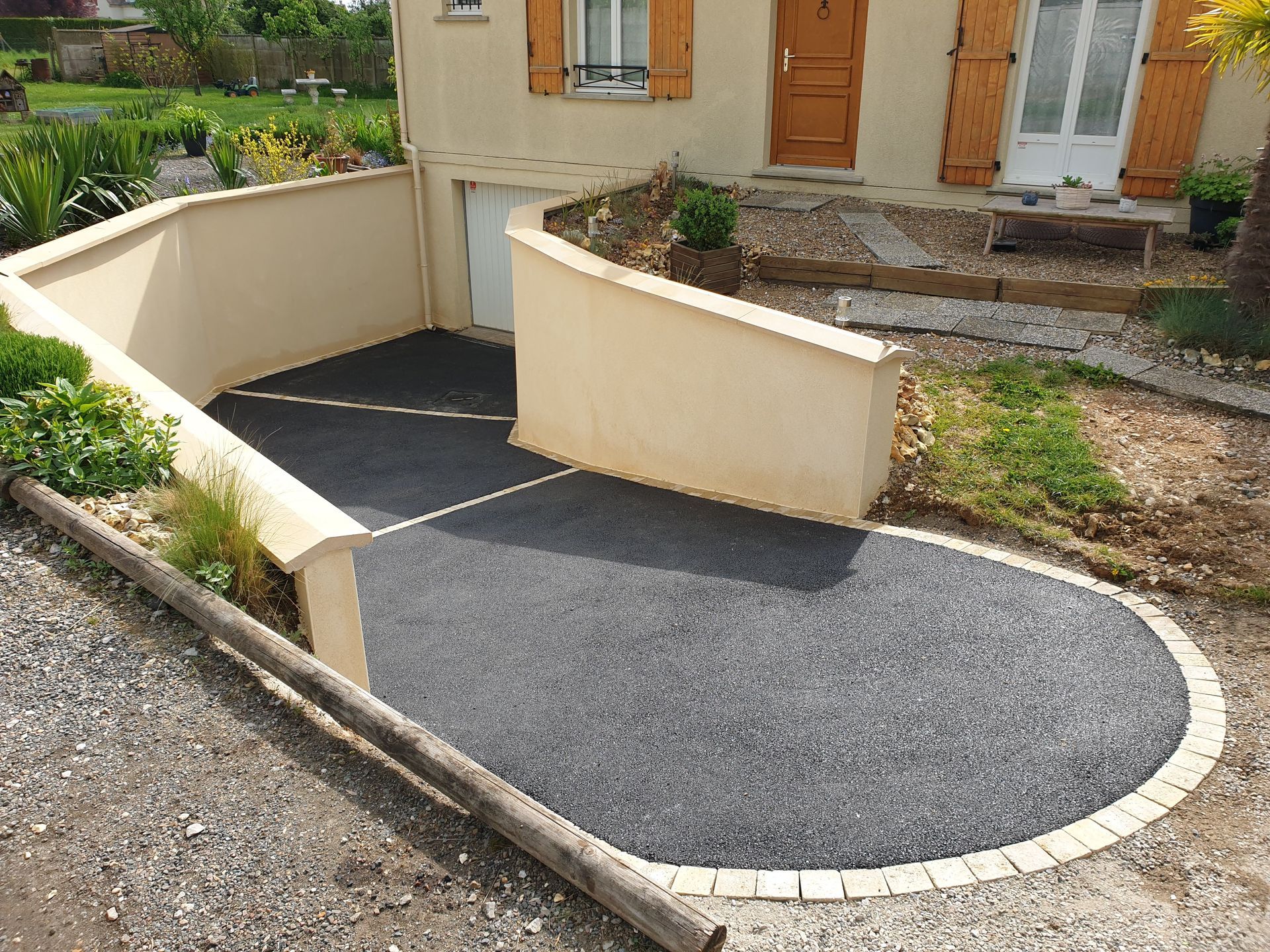 Une allée pavée grise descend en pente douce vers l'entrée d'un garage en sous-sol, bordée de murets en béton couleur crème.