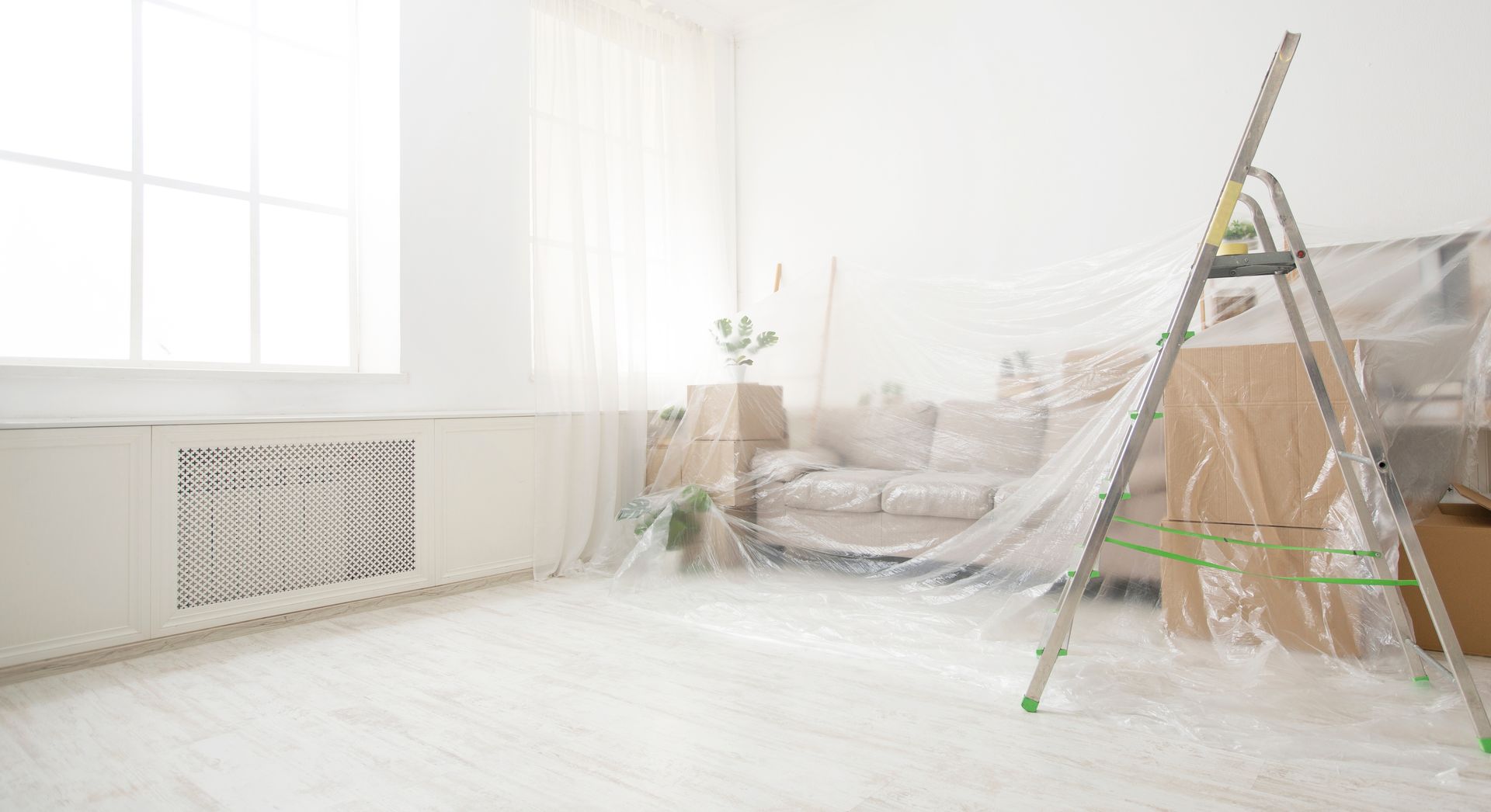 Une pièce lumineuse en cours de rénovation, avec un escabeau, des meubles recouverts de bâches en plastique et des murs blancs.