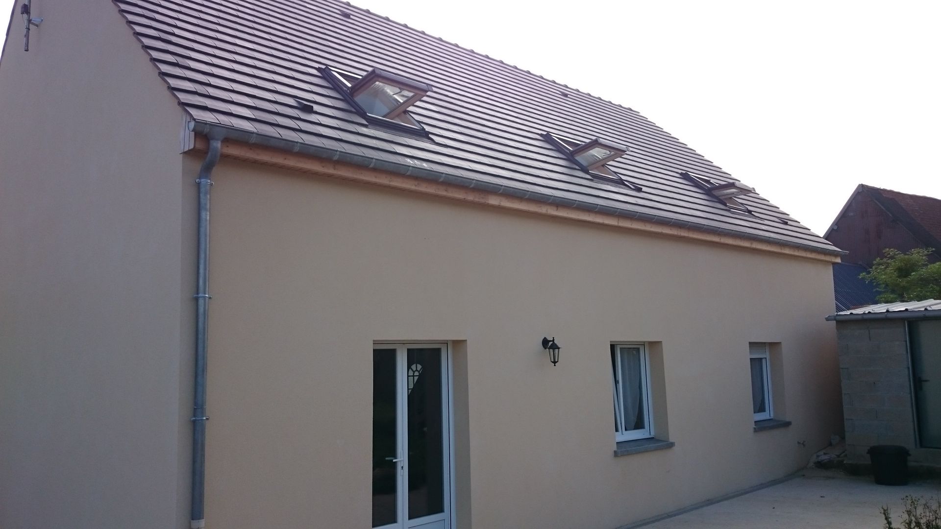 Une maison beige avec une porte vitrée et deux petites fenêtres et un toit de tuiles composé de trois puits de lumière.