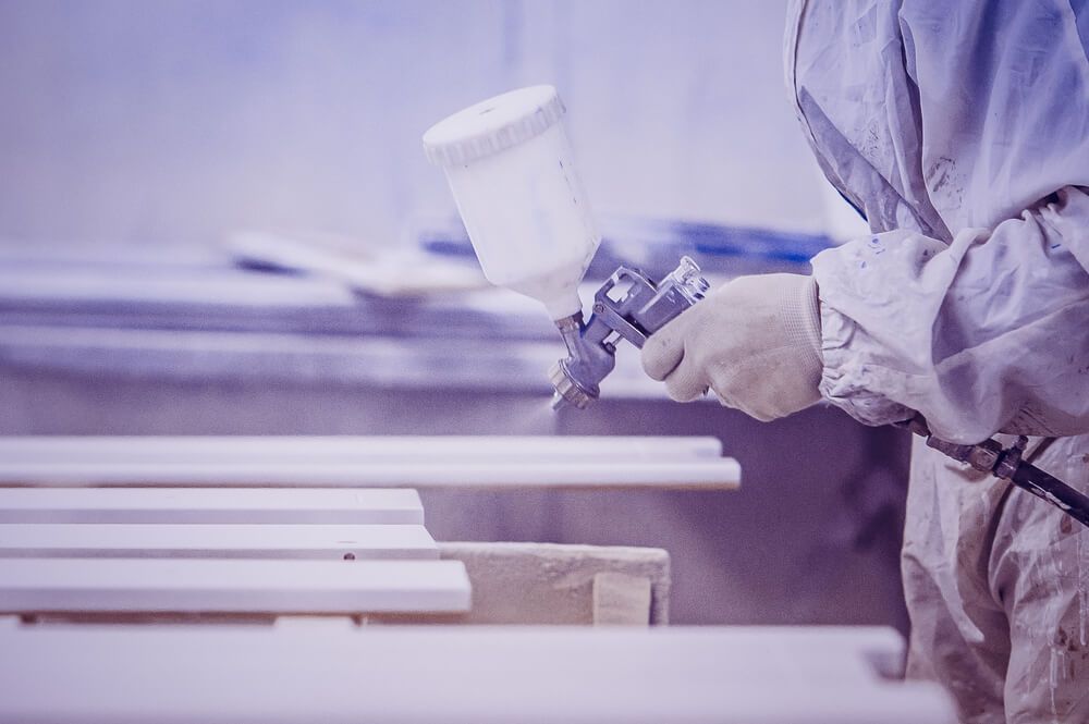 Une personne en combinaison de protection pulvérise de la peinture sur des morceaux de bois blanc à l'aide d'un pistolet à peinture.