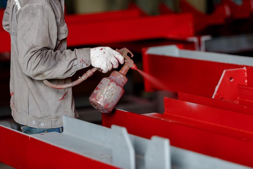 Une personne pulvérise de la peinture rouge sur des poutres métalliques.