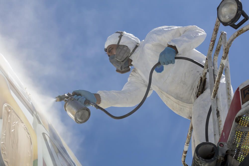 Un homme en combinaison de protection vaporise de la peinture sur une structure, sur fond de ciel bleu.