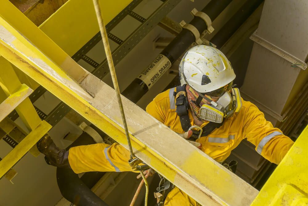Un ouvrier en combinaison jaune et masque respiratoire escalade une structure jaune, équipé d'un harnais de sécurité.