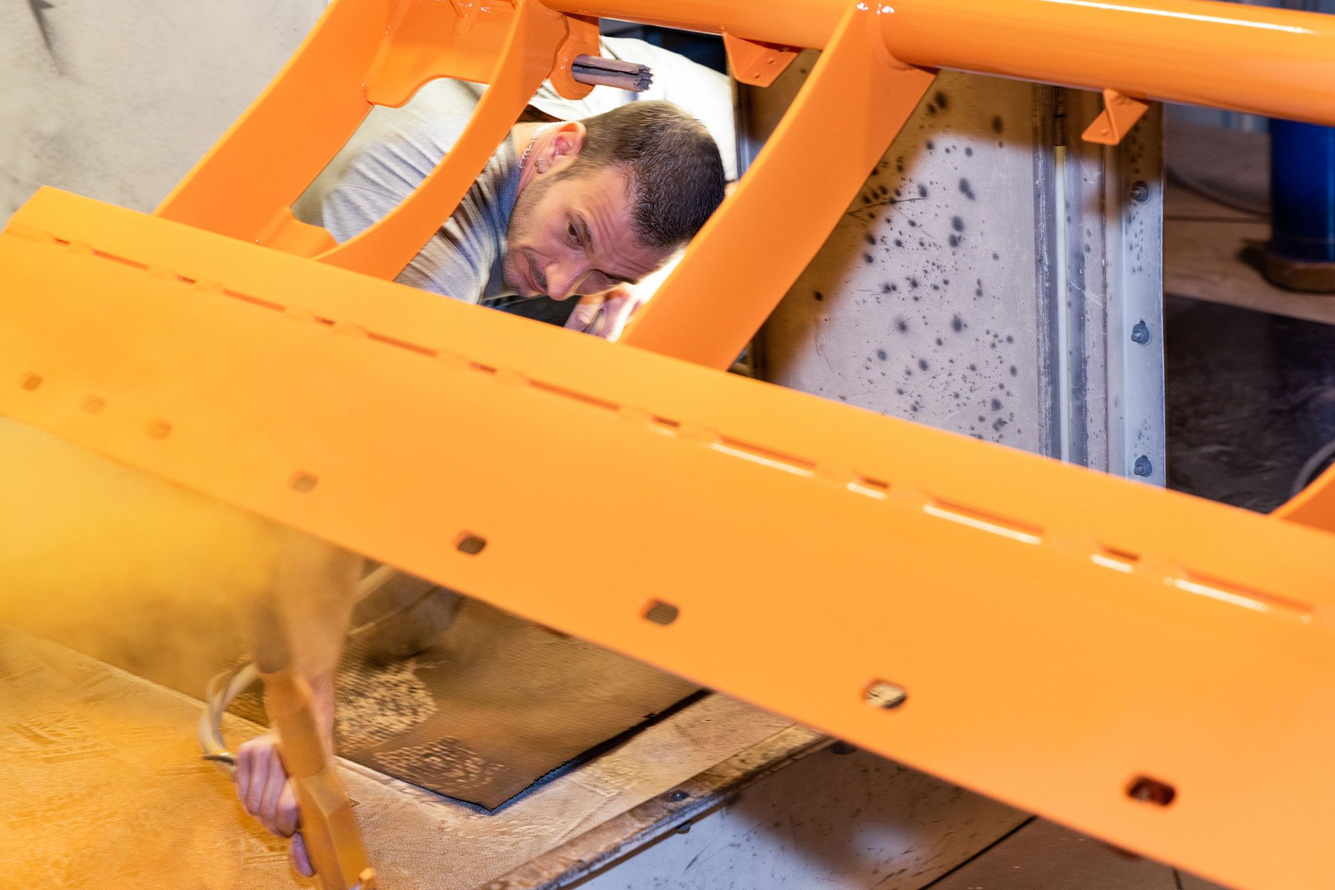 Un homme travaille sur une structure métallique orange dans un atelier, se penchant pour inspecter.