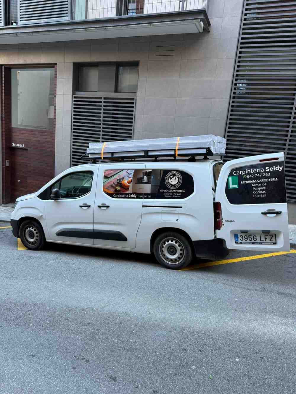 Furgoneta blanca de trabajo aparcada en una calle estrecha junto a un edificio, con escaleras en el techo y gráficos.
