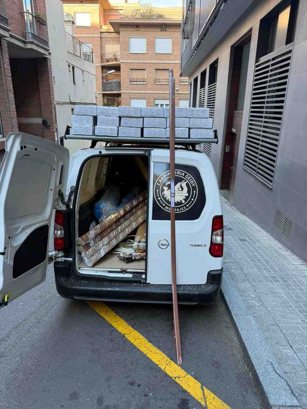 Furgoneta blanca en un callejón estrecho con las puertas traseras abiertas, cargada con largas tablas de madera.