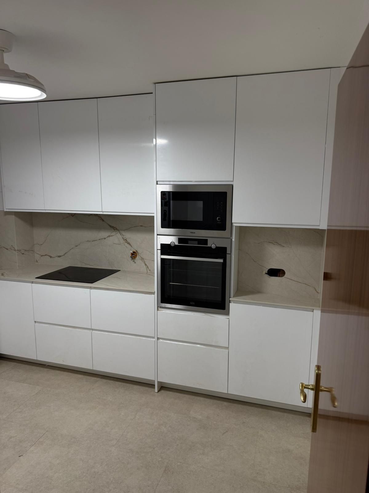 Cocina moderna blanca con horno empotrado, microondas y armarios blancos sobre paredes beige.