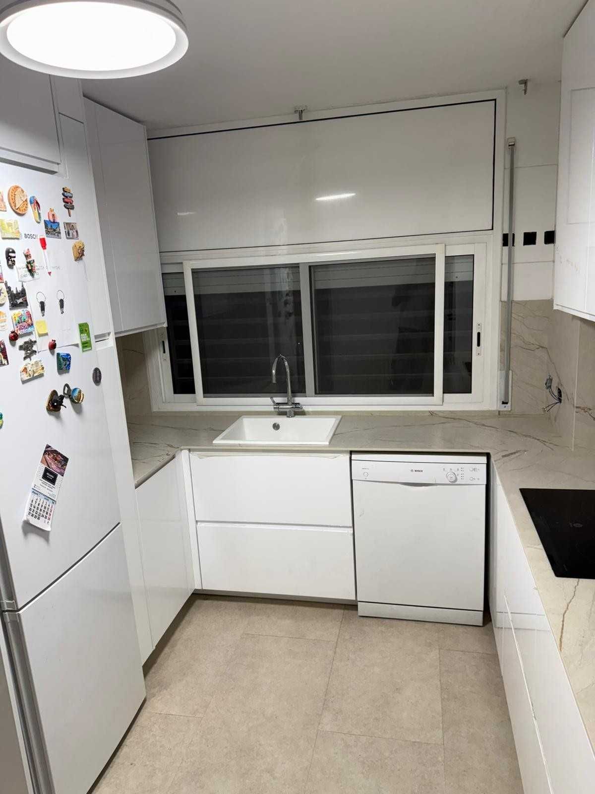 Pequeña cocina blanca con refrigerador, fregadero debajo de una ventana, lavavajillas y encimera negra.