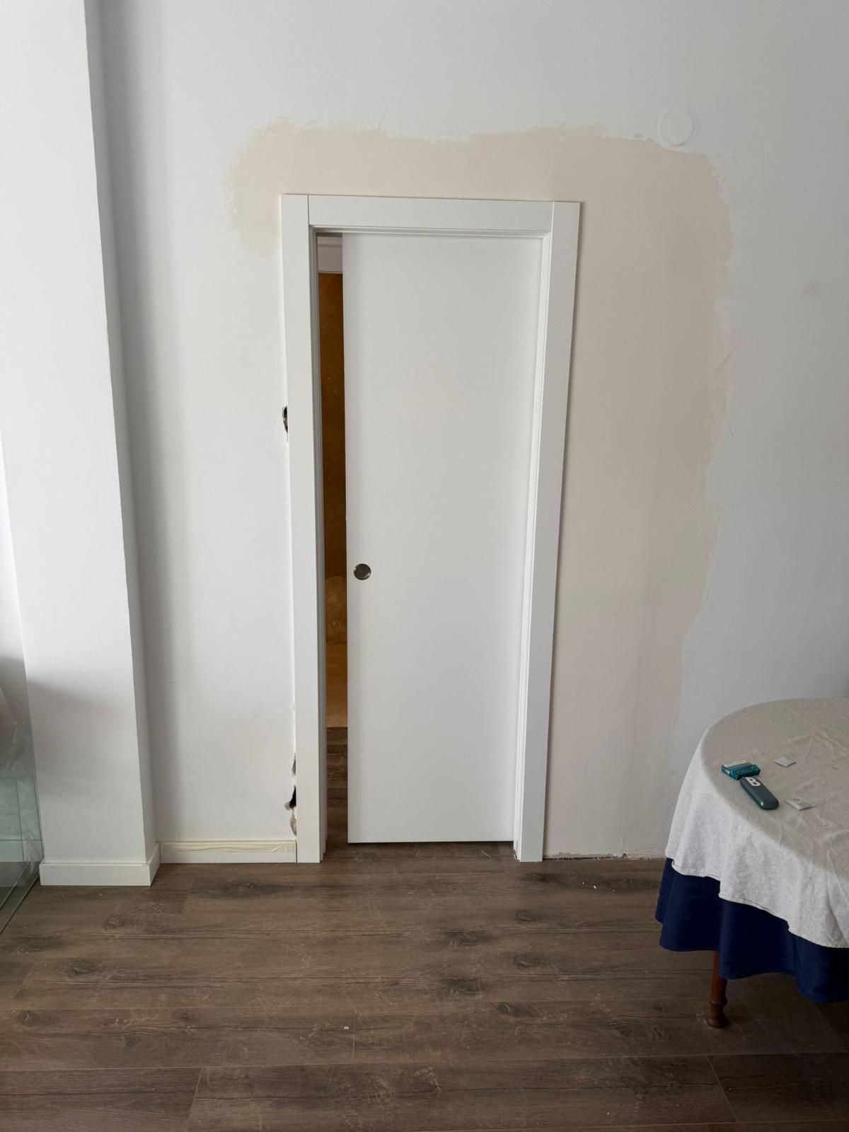 Una estrecha entrada en una pared blanca con una puerta blanca entreabierta, suelo de madera y una mesa a la derecha.