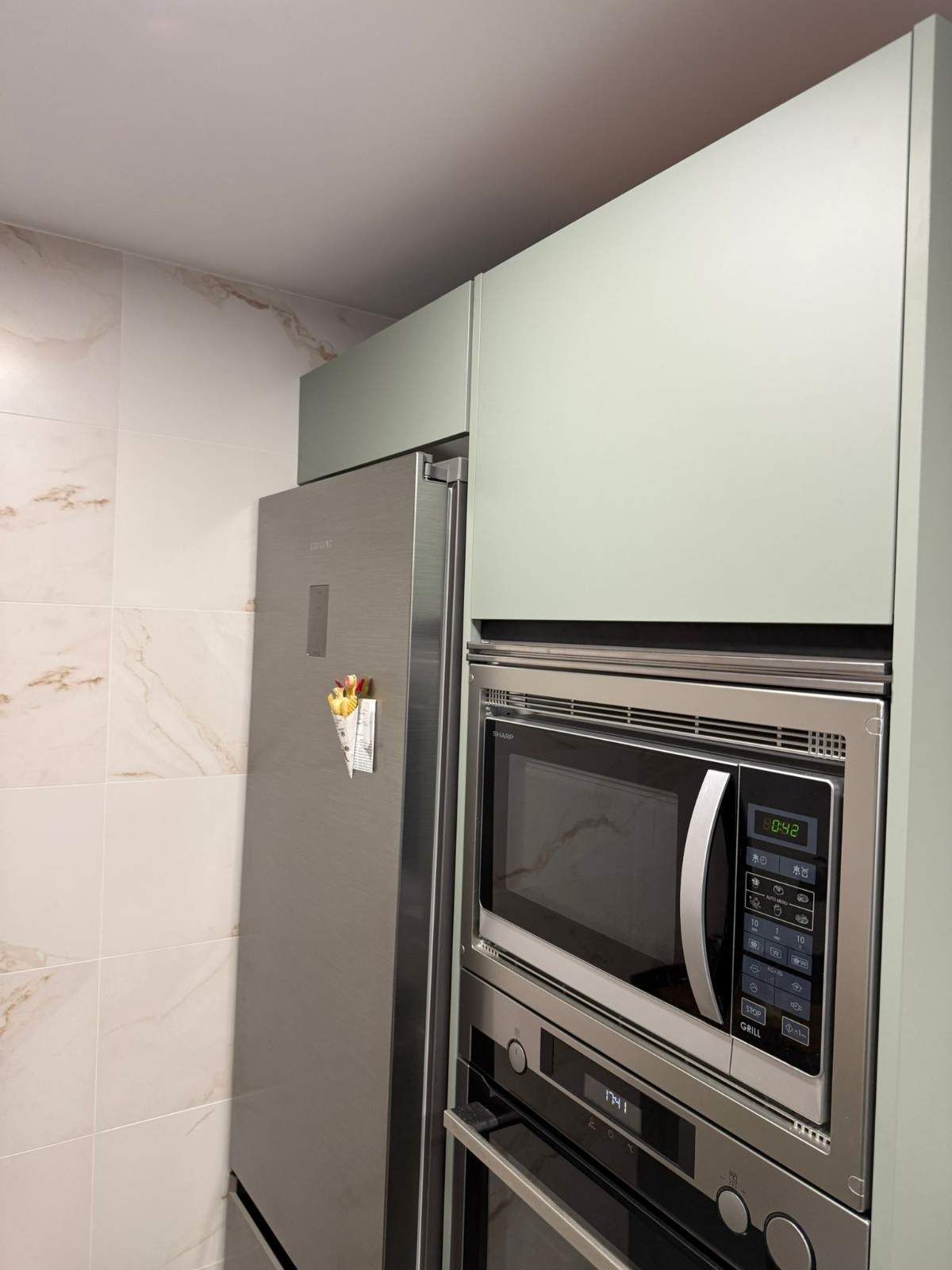 Rincón de cocina con refrigerador de acero inoxidable y microondas empotrado bajo gabinetes de color verde claro.