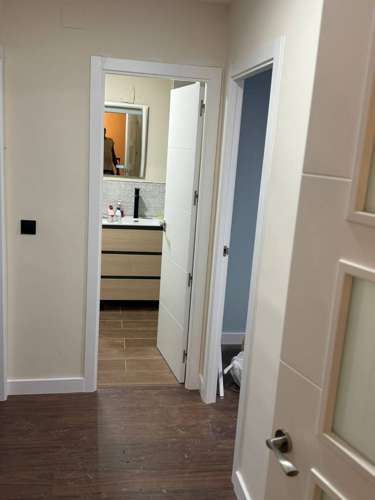 Pasillo con tocador de baño al frente, puertas blancas abiertas y suelo de madera oscura.