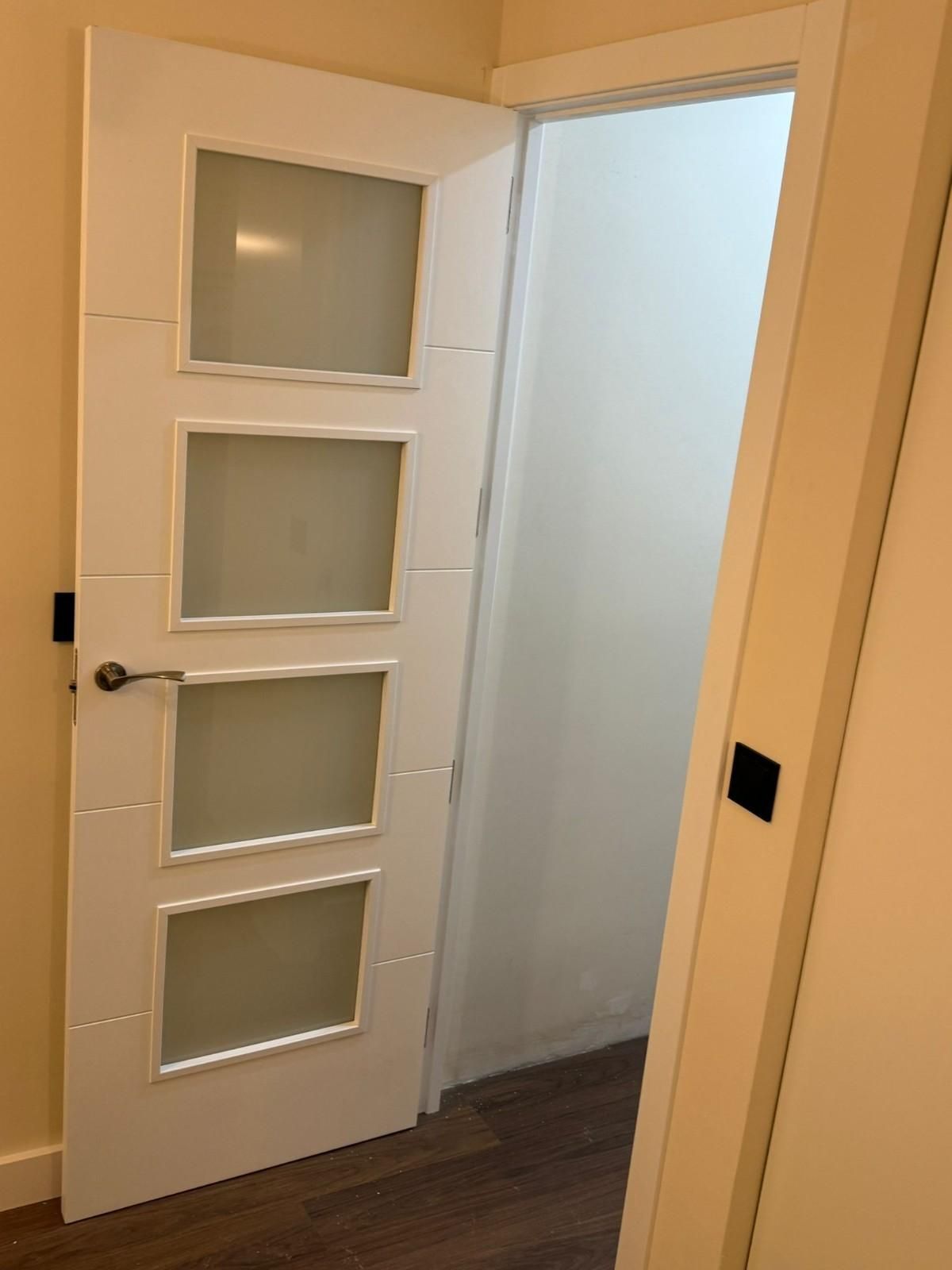 Se abre una puerta blanca con paneles de vidrio esmerilado, que deja ver un pasillo luminoso y vacío con suelo de madera.
