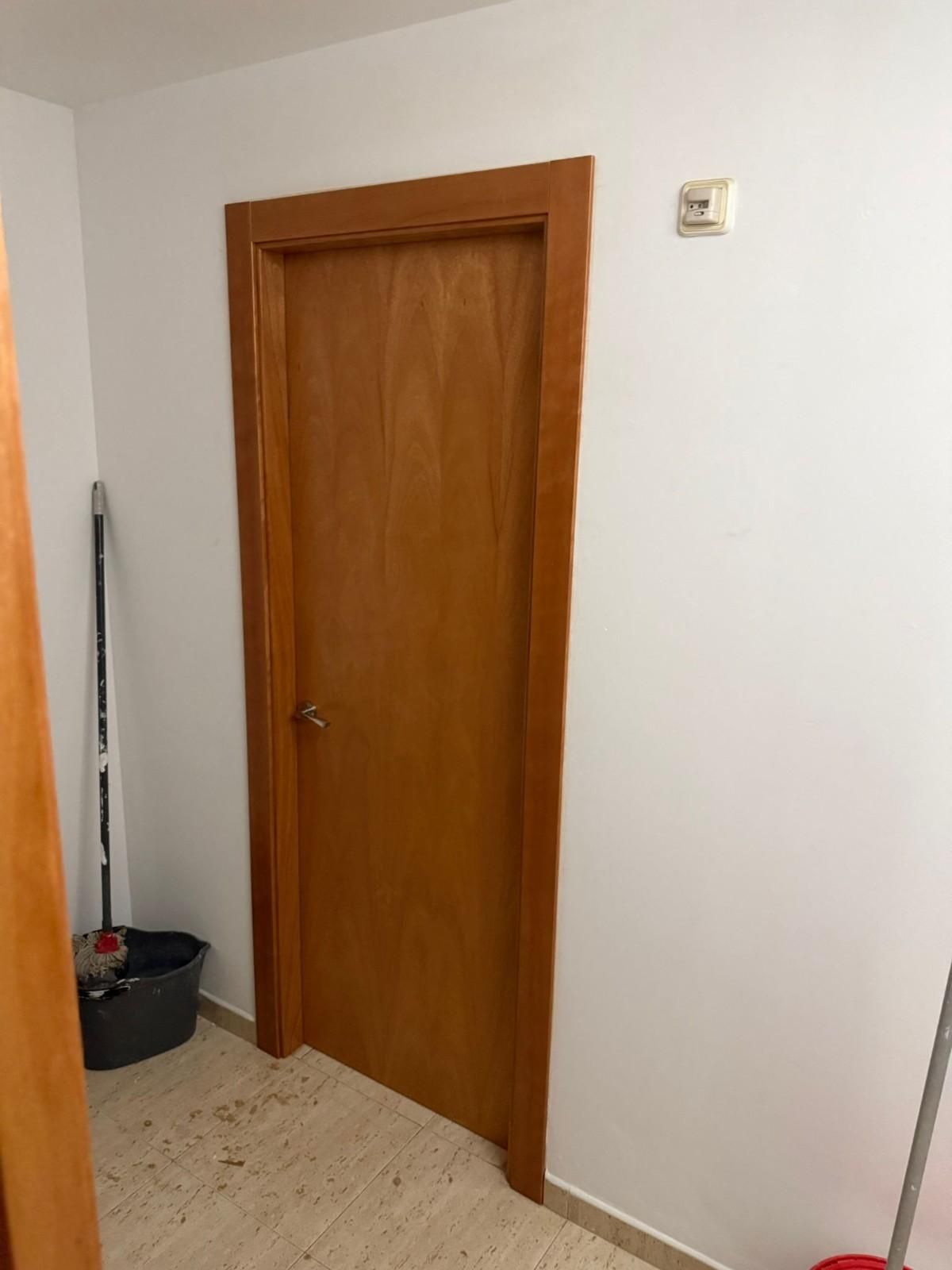 Puerta interior de madera en un pasillo blanco con suelo de baldosas beige y utensilios de limpieza a su lado.