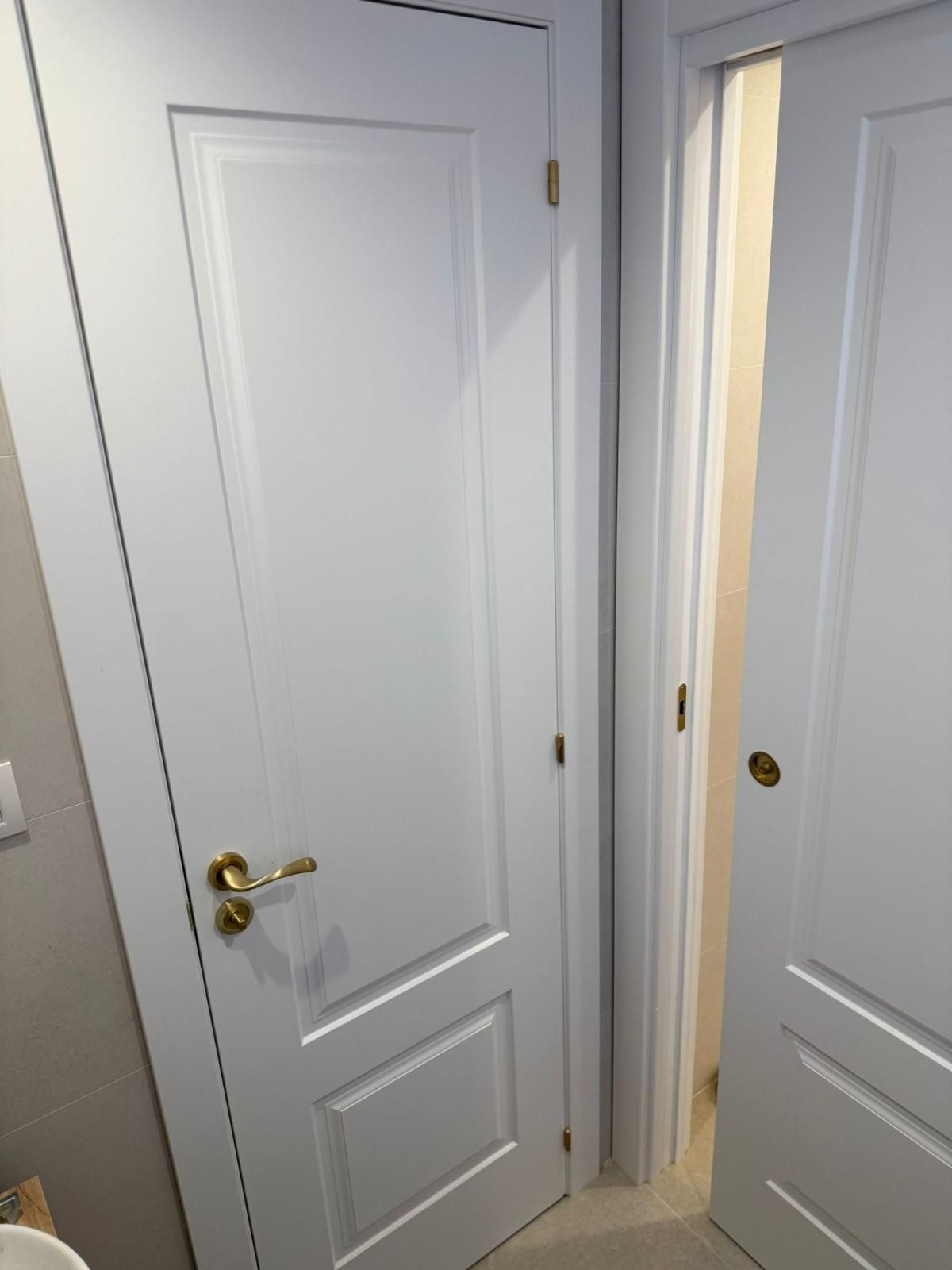Dos puertas interiores blancas en un pasillo estrecho, una de ellas ligeramente abierta por donde entra la luz.