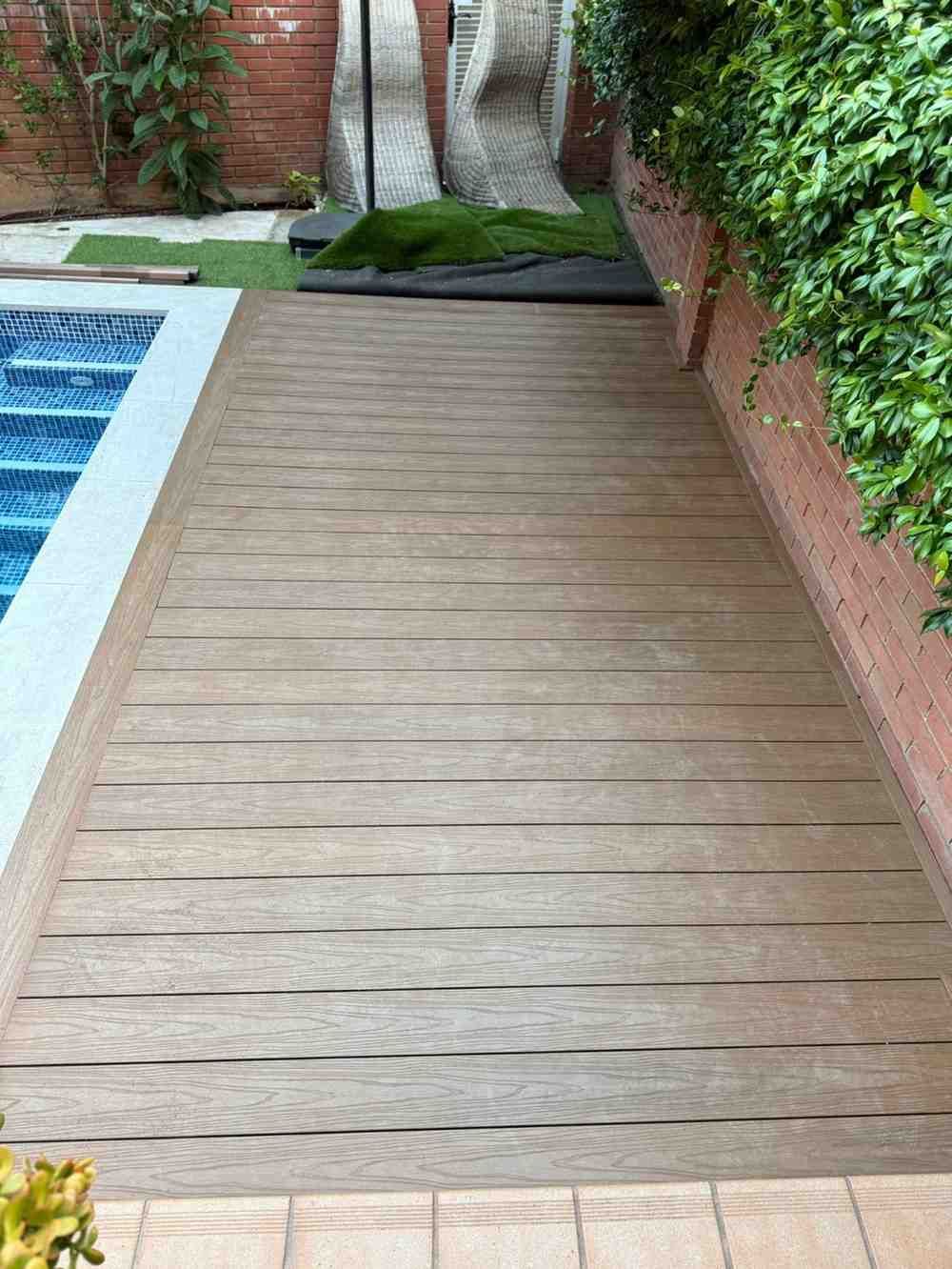 Terraza de madera junto a una piscina azul y un muro de ladrillo con vegetación.
