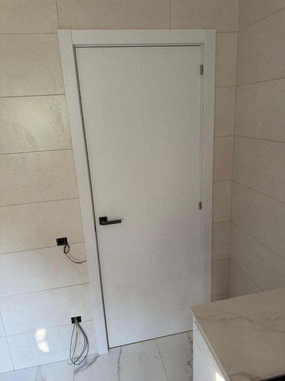 Puerta interior blanca en una habitación alicatada con tirador negro y toma de corriente en la pared cercana.