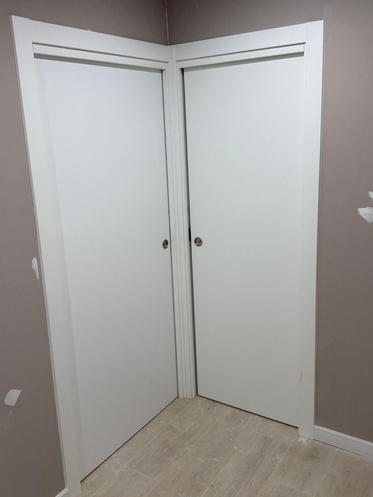 Puertas dobles blancas de armario en un rincón beige de una habitación vacía con suelo de madera clara.