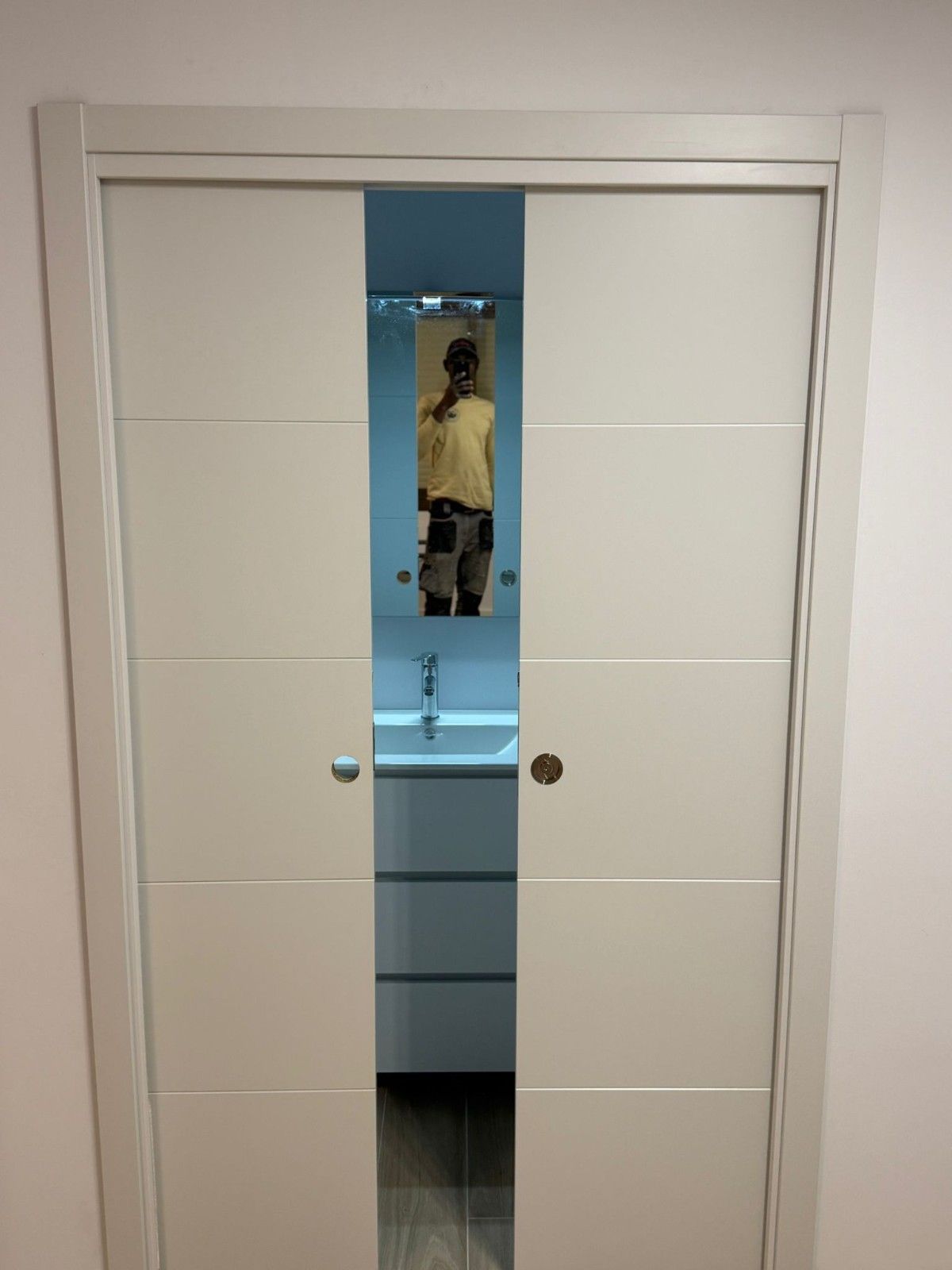 Puerta de baño blanca con una estrecha ventana vertical que deja ver un lavabo y un espejo en el interior.