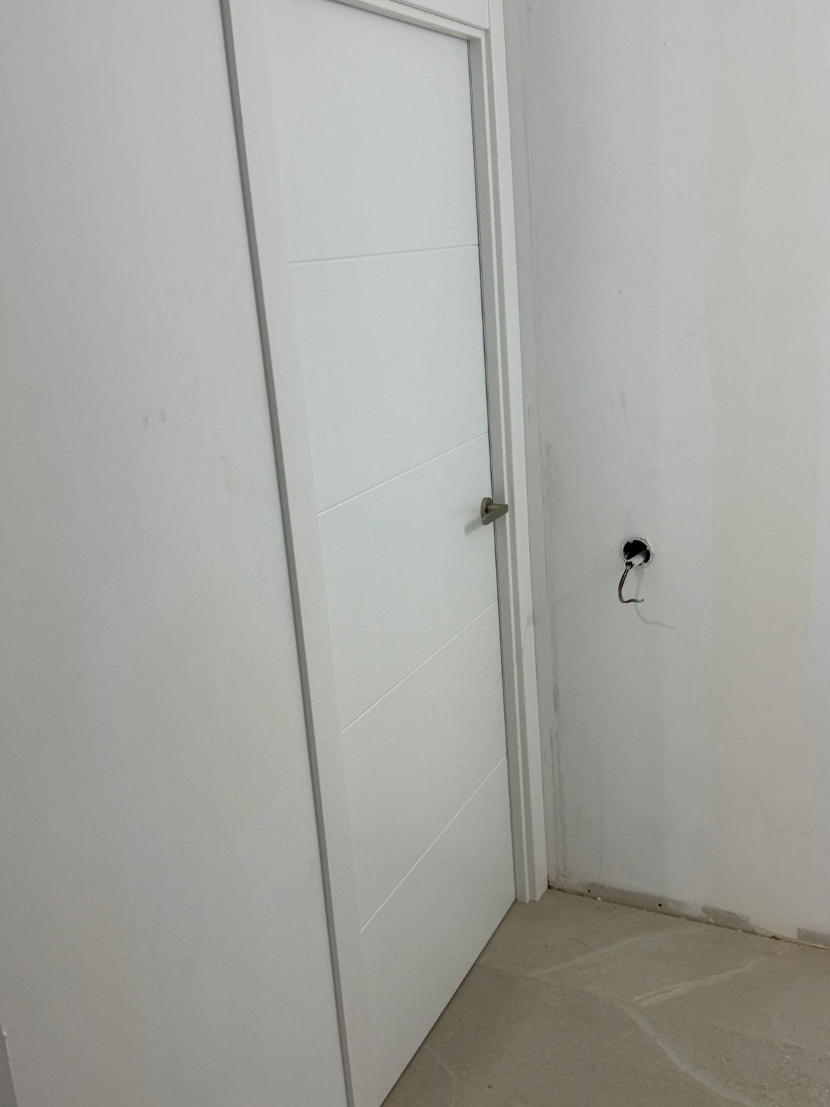 Puerta interior blanca junto a una pared blanca lisa con un enchufe eléctrico suelto y molduras de suelo sin acabado.