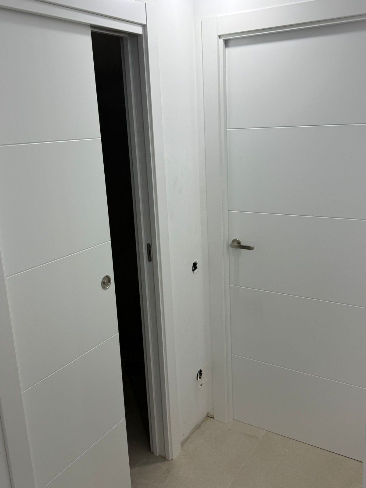 Pasillo blanco con dos puertas cerradas y una puerta ligeramente abierta que deja ver una habitación oscura.